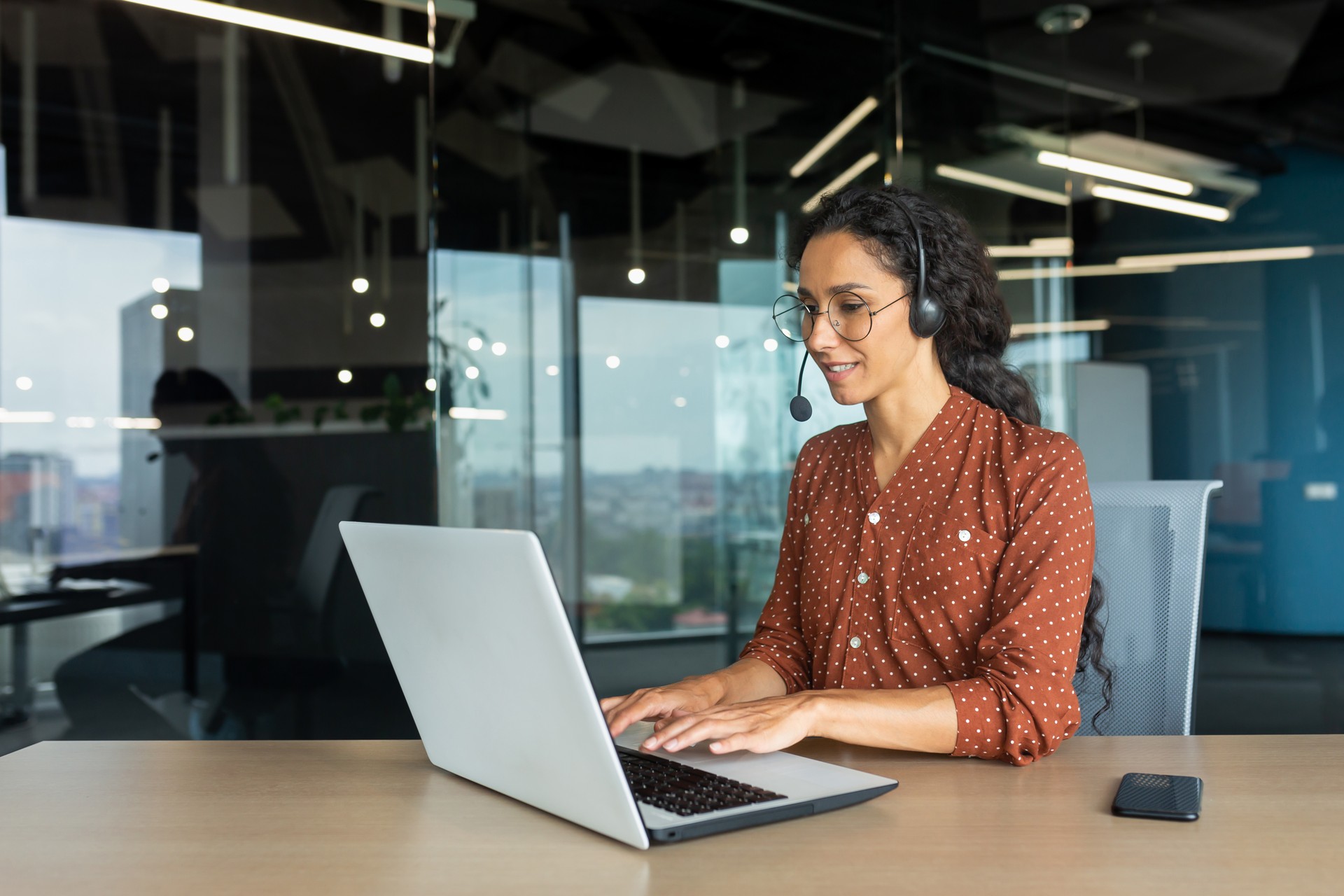 Smiling hispanic businesswoman working inside office with laptop and headset for video call, woman sitting at workplace happy working with clients